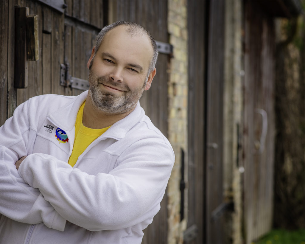 Malermeister Mike Schulz vor einer alten Holzscheune, freundlich lächelnd mit verschränkten Armen